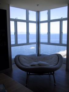 A képen egy fotel látható nagy üvegablak előtt rálátással a vízre.