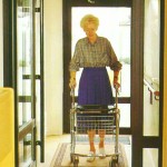 A képen a lépcsőházban egy kerekes kocsit toló idős néni látható.