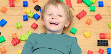 A képen egy kisfiú látható, amint mosolyogva legó kockák között fekszik.