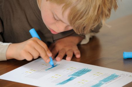 a képen egy down szindrómás kisfiú látható tanulás közben. egy kék filccel színez ki kockákat.