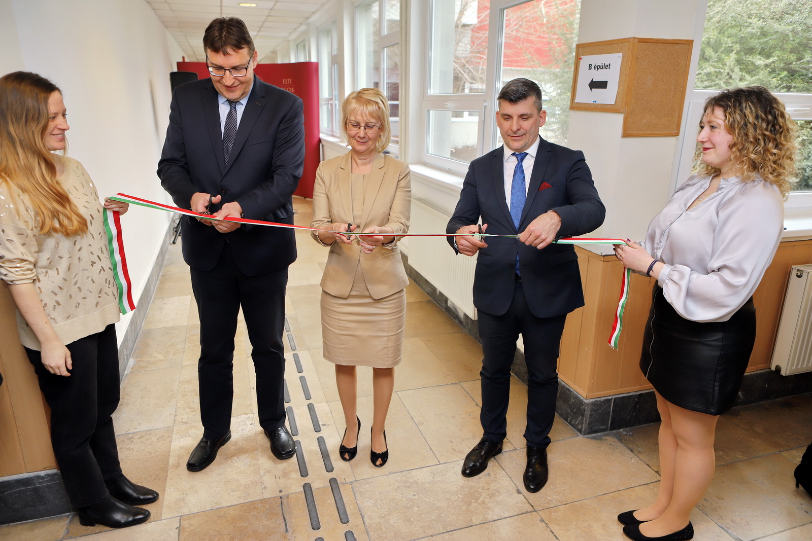 A képen egy magyar nemzeti színű szalagot vág át egy nő és két férfi. A szalagot balról és jobbról fiatal lányok tartják. A szalagátvágás egy folyosón történik.